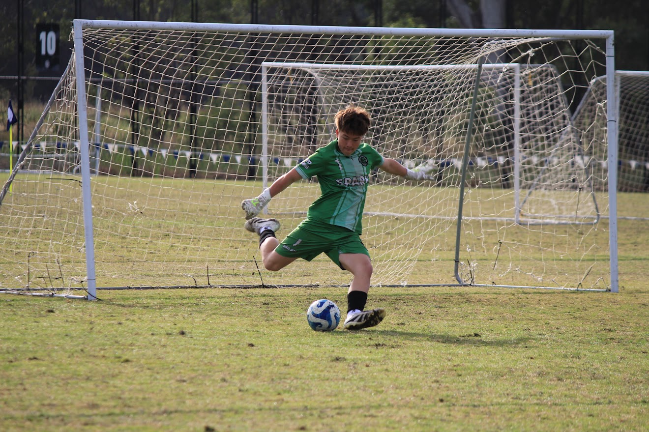  Goalkeeper in a green Spark Futbol kit kicking the ball out from the goal during a match on a grass field.